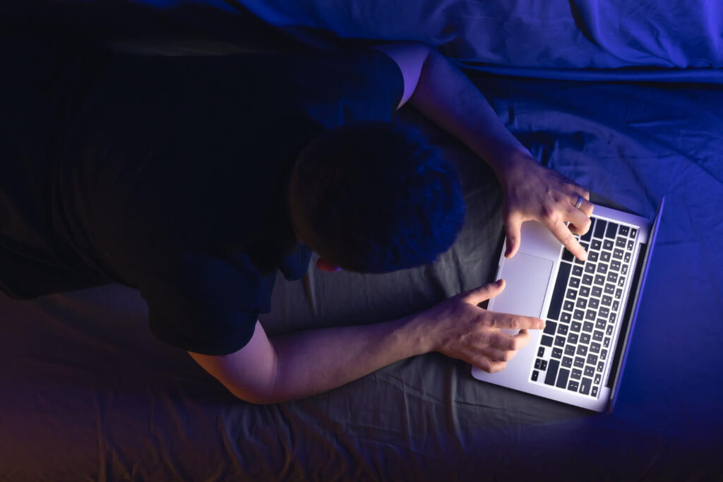Homem deitado na cama à noite, utilizando um notebook com iluminação azul em um ambiente escuro, representando o uso de telas antes de dormir e um hábito que prejudica a higiene do sono.