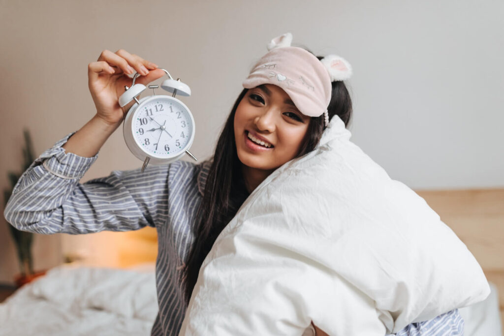Mulher jovem sorrindo ao acordar, segurando um despertador e abraçando um travesseiro, usando pijama e máscara de dormir, em um ambiente aconchegante, transmitindo a sensação de descanso, leveza e uma rotina de sono bem regulada.
