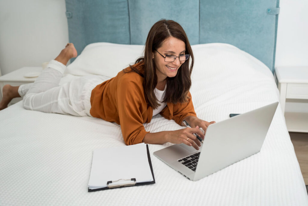 Mulher deitada na cama utilizando um notebook e anotando informações, representando o hábito de trabalhar na cama, que pode prejudicar a higiene do sono.