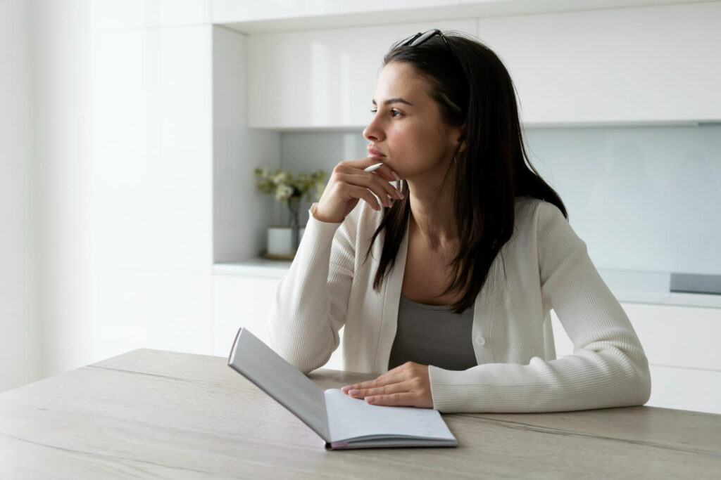 Mulher sentada à mesa com caderno aberto, olhando para o lado com expressão distraída, sugerindo dificuldade de concentração ou déficit de atenção.