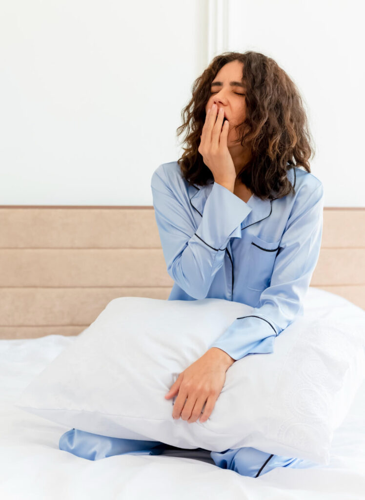 Mulher sentada na cama usando pijama, bocejando e com expressão de cansaço, segurando um travesseiro, sugerindo dificuldade para acordar ou noite mal dormida.