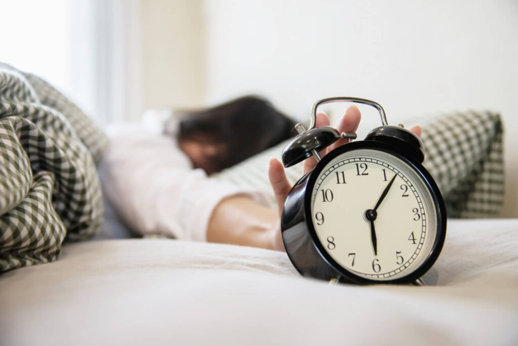 Pessoa com dificuldade para acordar ao lado de um despertador, representando a relação entre sono irregular e pressão alta e o que fazer para melhorar a qualidade do descanso.