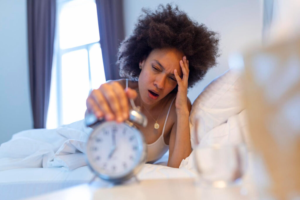 Mulher acordando cansada e estressada na cama ao desligar o despertador pela manhã, demonstrando os efeitos da privação do sono no cansaço físico e mental.