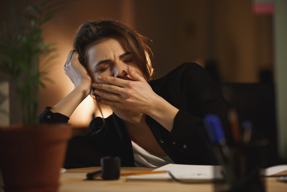 Mulher bocejando e demonstrando cansaço excessivo durante a noite, ilustrando os efeitos da desregulação do ritmo circadiano e da privação do sono na rotina e na produtividade.