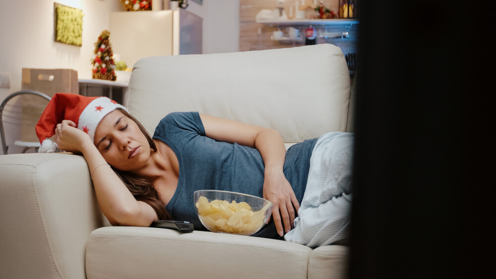 Mulher dormindo no sofá enquanto assiste televisão e consome alimentos ultraprocessados à noite, ilustrando hábitos de higiene do sono inadequados que podem contribuir para a privação do sono.